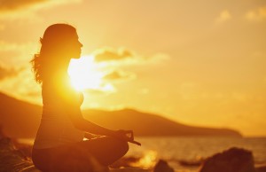 A pregnant person is meditating on the beach with the sunset in the background.